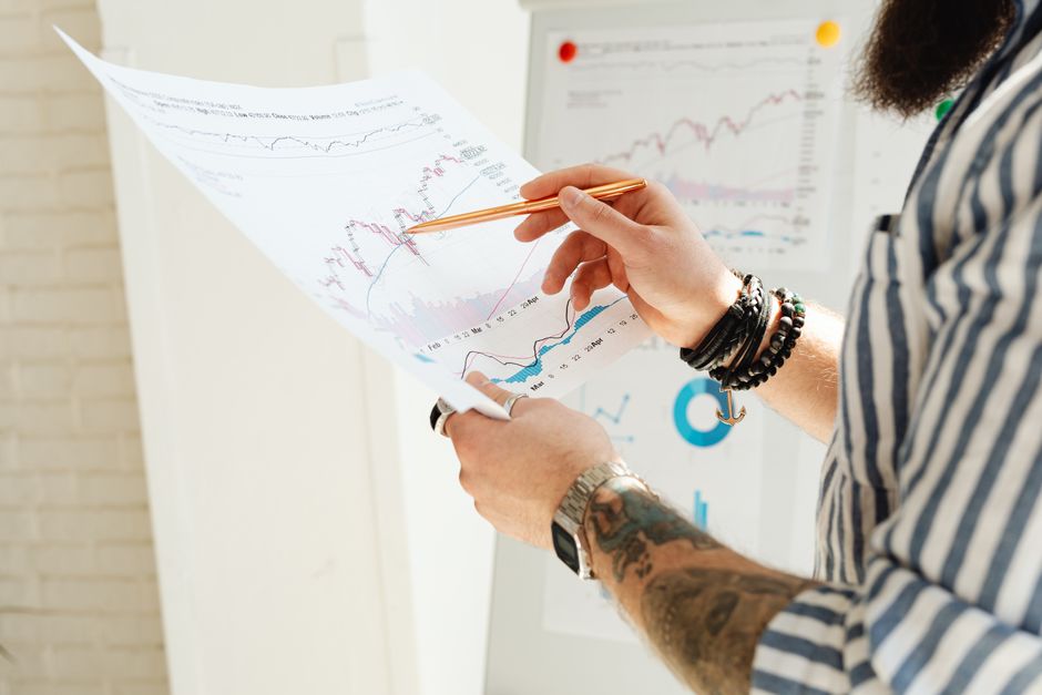 Diversidade nas Empresas — A man analyzing stock market charts with a pen, holding a paper report indoors.